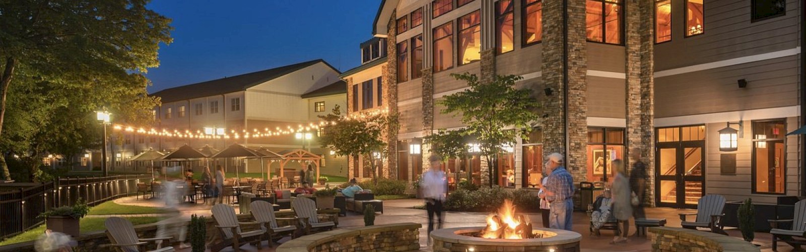 An evening scene showing a resort with a fire pit, string lights, Adirondack chairs, and a lodge-style building with large windows and people relaxing around.