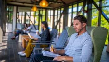 People are sitting outdoors in a shaded seating area, some working on laptops or taking notes, enjoying a peaceful environment.