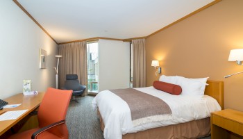 A modern hotel room featuring a bed with a bolster pillow, a desk with an orange chair, a cozy lounge chair, and large windows with curtains.