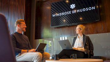 Two professionals have a meeting in a modern office, smiling and working on their laptops, with a screen showing 