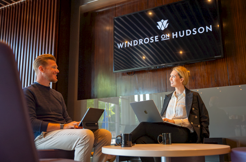 Two professionals have a meeting in a modern office, smiling and working on their laptops, with a screen showing 