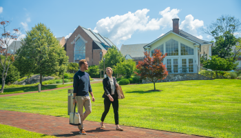Two people walk and talk on a brick path in a lush park, with modern houses and bright blue sky in the background.
