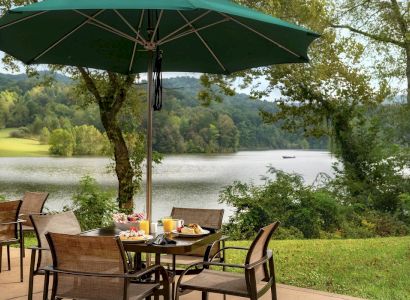 An outdoor dining setup by a lake with a green umbrella, surrounded by nature. There are plates of food and drinks on the table, with scenic views.