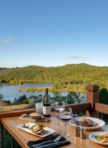 A table set with food, wine, and glasses overlooks a scenic lake surrounded by forests and hills, under a clear blue sky.