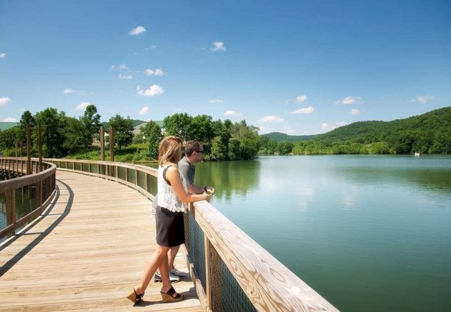 Two people are standing on a wooden boardwalk overlooking a serene lake with green hills and trees in the background on a clear day.