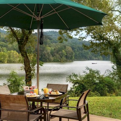An outdoor dining area with a table set for breakfast, under a large green umbrella, overlooking a scenic lake and surrounded by trees.
