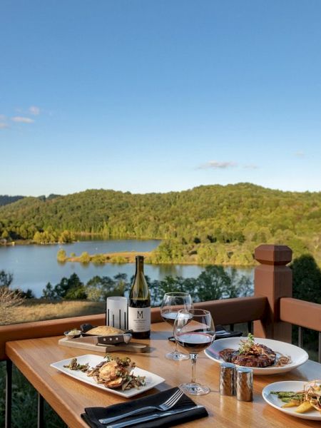 A dining table with plates of food, wine, and glasses, set on a balcony overlooking a scenic lake and forested hills under a clear blue sky.