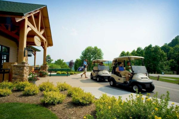 Two golf carts are parked outside a building with one person sitting in each cart, while another person walks away carrying golf clubs.