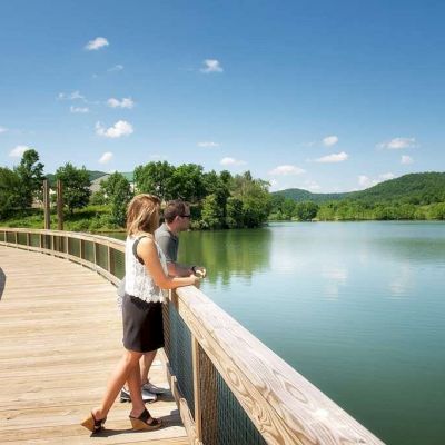 A couple is standing on a wooden boardwalk, overlooking a peaceful lake surrounded by green mountains and clear, blue skies.