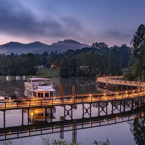 A serene landscape featuring a wooden pier winding over calm water, a moored boat, surrounded by trees and distant mountains under a twilight sky.
