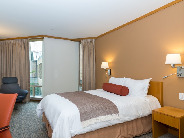 A comfortable hotel room located inside Croton Hall showing a bed, modern chair, lamp, and a window overlooking greenery.