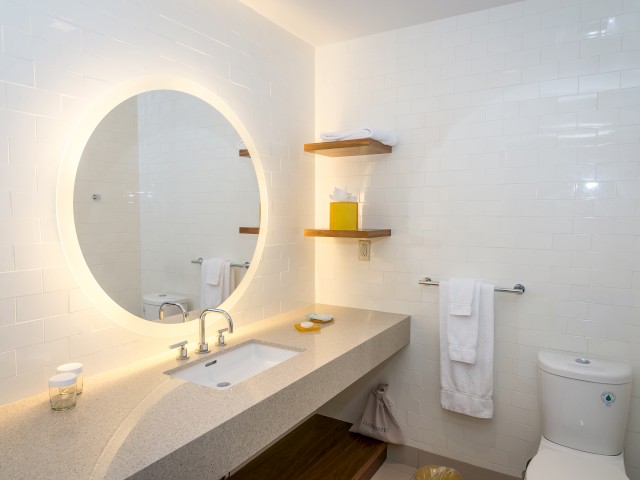 The image shows a modern bathroom in The Village accommodations with a circular, backlit mirror above a large countertop sink, white towels, shelves, and a toilet.