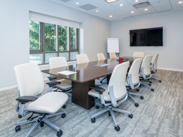 The image shows a modern conference or board room with a long table, white ergonomic chairs, a wall-mounted TV, large a window overlooking greenery.