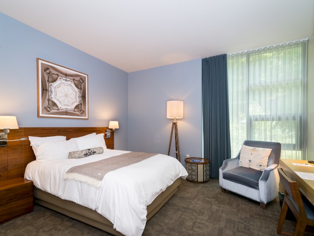 A modern hotel room inside The Village with a bed, two bedside tables with lamps, framed artwork, a comfortable armchair, a floor lamp, and a window overlooking greenery.