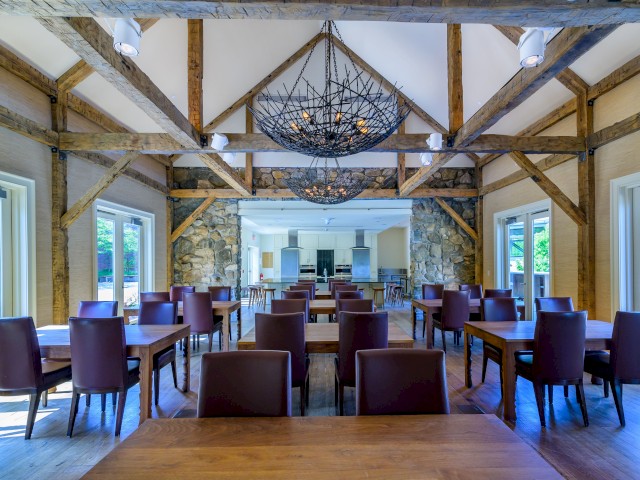 Picture shows the dinning area inside the Carriage House a spacious, rustically elegant room with high ceilings, wooden beams, tables, leather chairs, and large windows, featuring unique chandeliers.