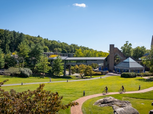 A scenic view of expansive green lawns with modern buildings like The Bridge elevated walkway connecting Croton Hall and The Village in the background. Surrounded by lush greenery, pathways, and people walking and cycling under a clear blue sky.