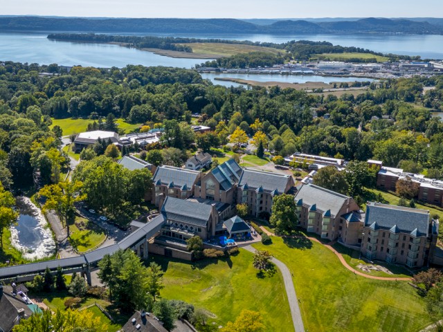 A scenic aerial view of the Windrose on Hudson on their 62-acre campus with multiple modern buildings, surrounded by lush greenery and a view of the Hudson River in the background.
