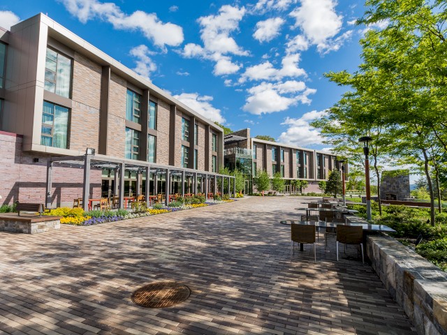 Image shows large brick patio outside The Village and River Fox. A modern building with brick walkways, outdoor seating, and lush greenery under a blue sky with scattered clouds.