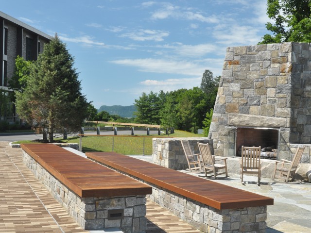 The image shows River Fox Commons, an outdoor area with wooden benches, a large stone fireplace, and wooden rocking chairs, surrounded by greenery and a building on the left.