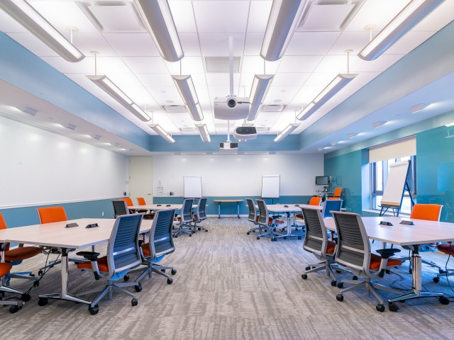 A modern conference room with multiple tables, orange ergonomic chairs, two whiteboards, and projection screens under bright ceiling lights with bright teal walls.