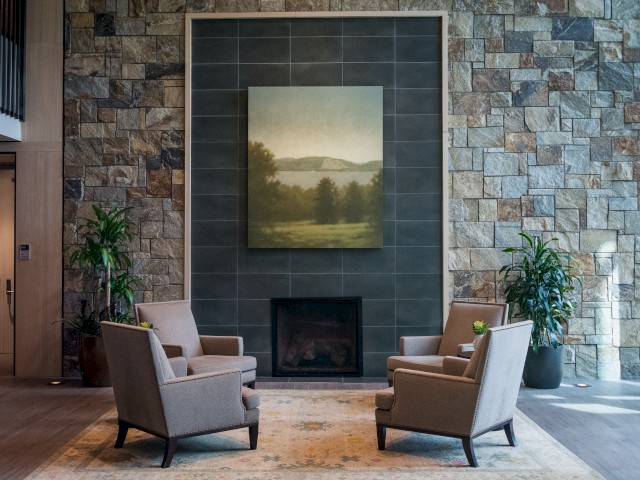 Picture shows The Village lobby cozy seating area with four armchairs, a fireplace, a large landscape painting, plants, and stone walls creating a warm welcoming ambiance.