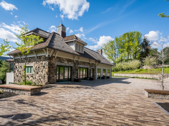 Image shows the Carriage House a stone building with large windows and a paved courtyard surrounded by trees under a blue sky with clouds. This building serves as a teaching kitchen and additional dining area.
