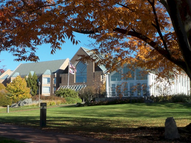 Croton Hall, the main building on property, a large glass front, red brick walls, and peaked roofs is surrounded by autumn trees, with a clear sky and well-maintained lawn in front.