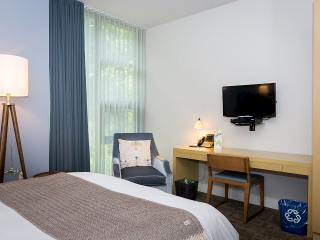 This image shows a modern hotel room inside The Village featuring a bed, armchair, desk with a chair, desk lamp, and mounted television.