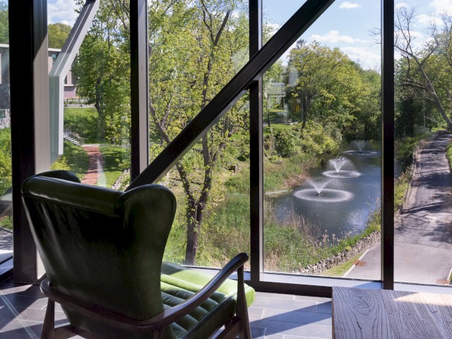 This image shows a climate controlled walkway connecting Croton Hall and The Village known as The Bridge. It shows a green chair facing large windows with a serene view of a lush green landscape with fountains in a stream..