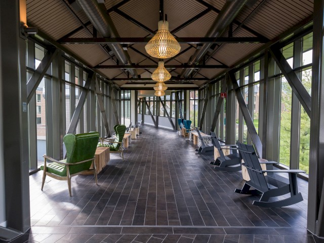 Image shows a walkway connecting Croton Hall and The Village known as The Bridge. A well-lit, glass-enclosed corridor with seating areas, featuring green and blue chairs, wooden tables, and elegant hanging lights.