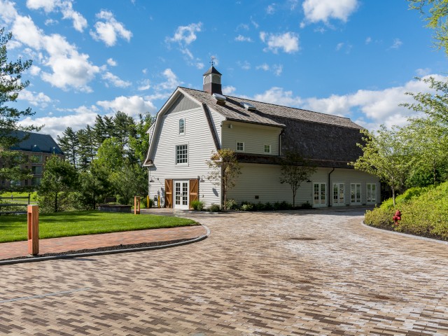 This image shows The Barn a large white farmhouse with a brick driveway, surrounded by lush greenery under a bright blue sky. This building features casual meeting and event space.