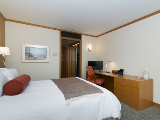 A comfortable hotel room located inside Croton Hall with a bed, desk, chair, computer, lamp, phone, wall art, and a door to a bathroom.