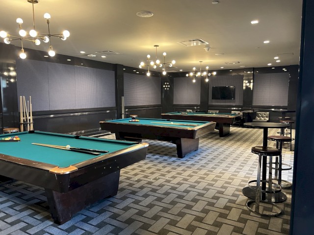 The image shows a modern game room with multiple pool tables inside the River Fox Restaurant and Bar. It shows retro overhead lighting fixtures, a checkerboard-patterned carpet, and bar-height tables.