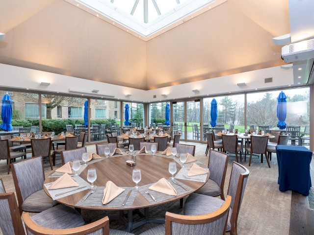 The spacious, well-lit Windrose Dining Room area with large windows, neatly set tables with glassware and napkins, and blue umbrellas with patio seating are visible outside.