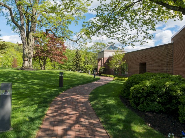 A brick walkway leading to Leadership Exchange curves through a green lawn with bushes, trees, through the expansive 62 acre campus.