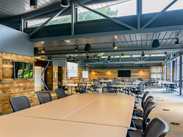 This image shows the Green House modern conference space with wooden walls, long tables, high industrial ceilings painted black with skylights. There are ergonomic chairs, and large screens, this space is powered entirely by green energy and built with recycled materials.