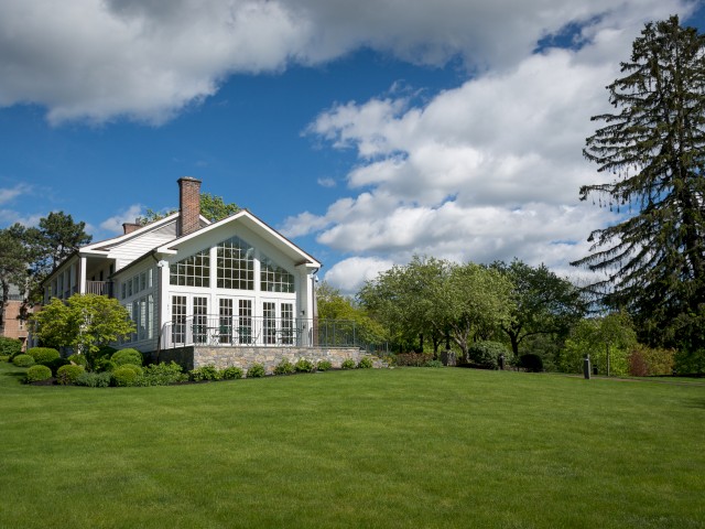 The image shows Cafe 56, a large historic white house with large windows, surrounded by lush greenery, a manicured lawn, and trees under a partly cloudy sky. Cafe 56 is the on-property coffee shop.