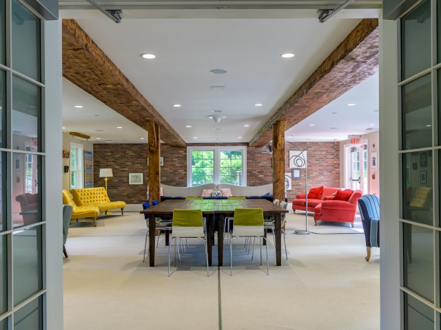 This image shows the communal space in The Barn featuring an open space colorful couches and large center table. Large windows overlook greenery.