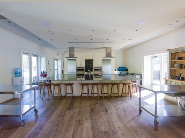 This image shows the inside of the Carriage House a modern kitchen with a large island, barstools, stainless steel appliances, and ample counter space in a bright, spacious setting. This space is as a teaching kitchen and additional dining area.