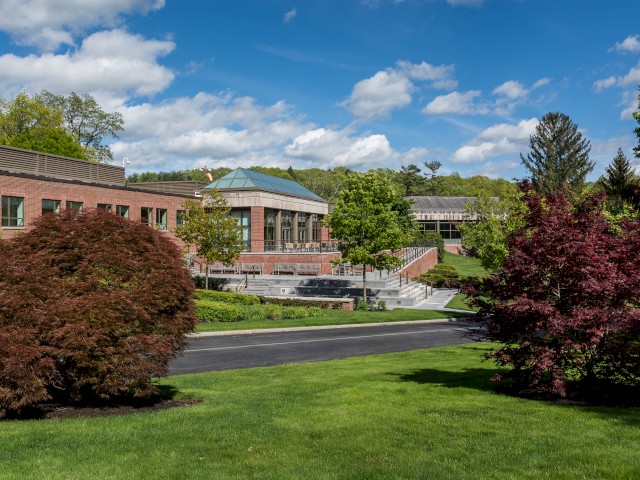 A scenic view of the Leadership Exchange a red-brick building, with a large patio for hosting events, green lawns, bushes, trees, and a clear blue sky with scattered clouds in the background.