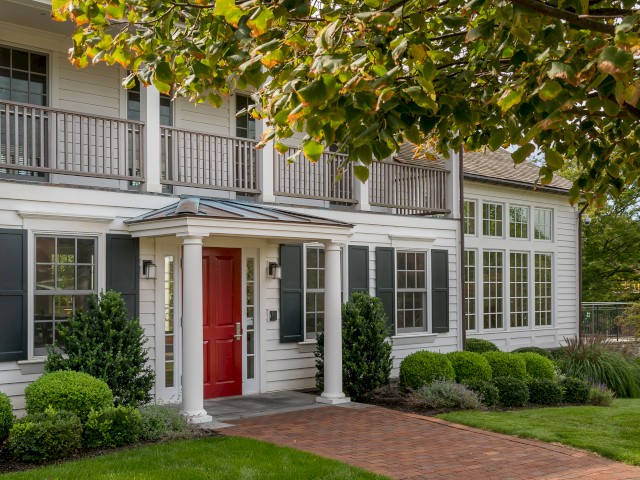 Image shows entrance to Cafe 56, a two-story historic house and the on-property coffee shop. Image shows a red door, white siding, black shutters, and manicured shrubs. A paved pathway leads to the entrance, surrounded by greenery.