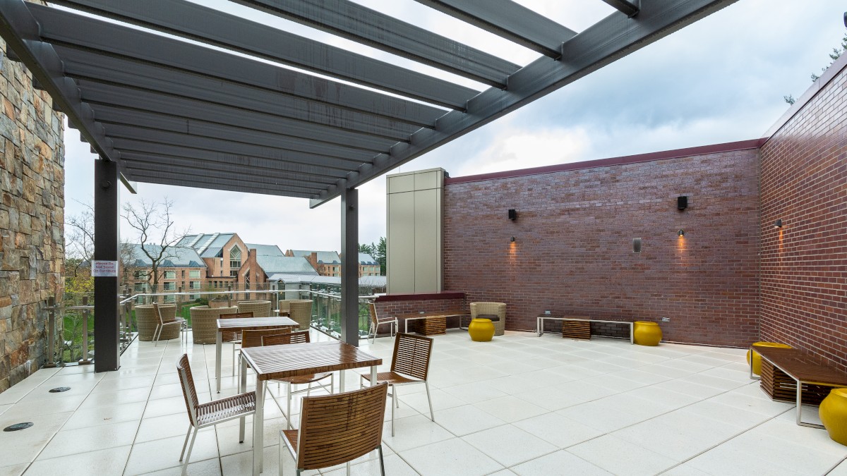 An outdoor patio area with pergola, wooden chairs, and metal tables, surrounded by brick walls and overlooking a scenic background with buildings in the distance.
