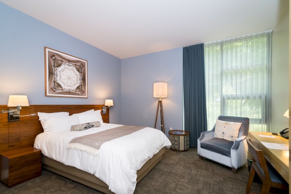 A modern bedroom with a double bed, wall art, large window with curtains, armchair, bedside tables, and floor lamp, featuring neutral colors.