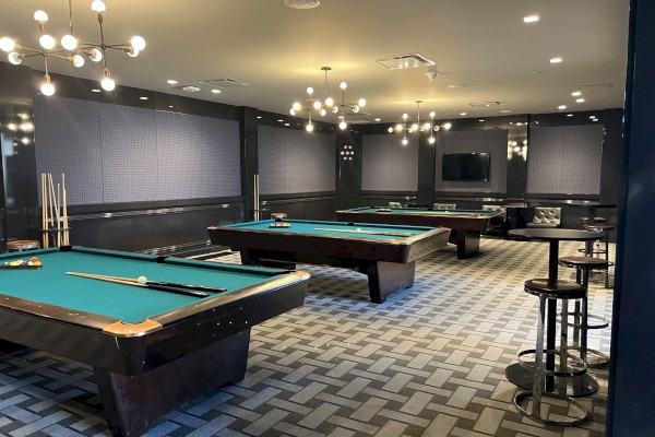 The image shows a stylish billiards room with several pool tables, modern light fixtures, stools, and a flat-screen TV mounted on the wall.