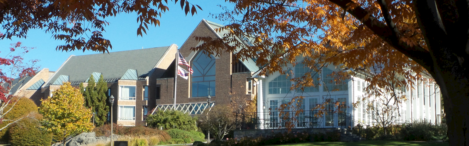 A building complex with peaked roofs, surrounded by trees with autumn foliage, and an open grassy area in the foreground, under a clear sky.