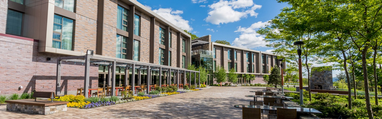 The image shows a modern building with large windows, outdoor seating, and a paved walkway. Trees and greenery line the area under a blue sky.