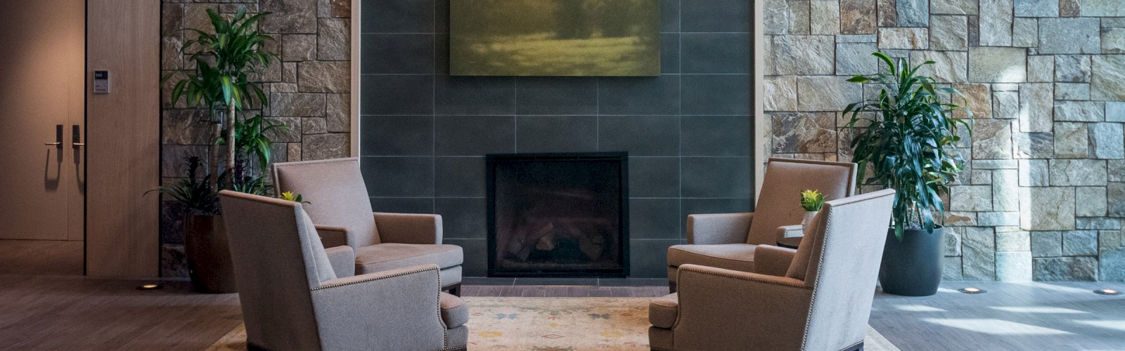 A cozy seating area with four armchairs, a rug, and a central fireplace with a landscape painting above it. The backdrop is a stone wall.
