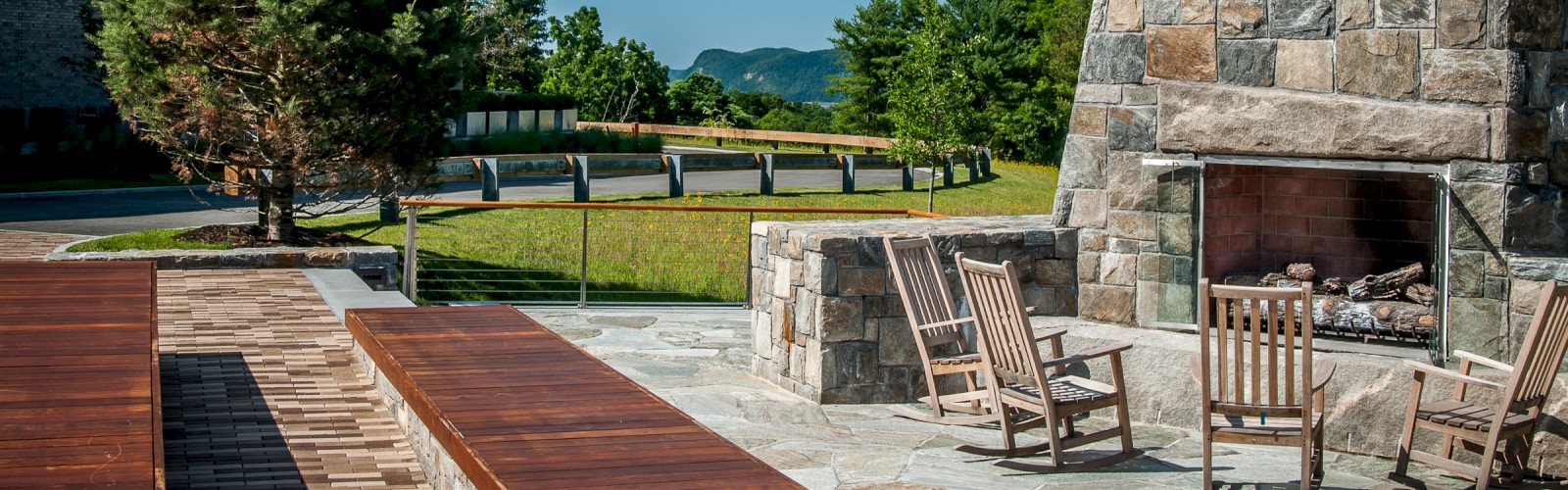 An outdoor patio has wooden benches, stone fireplace, and rocking chairs, surrounded by green trees and a blue sky in the background.