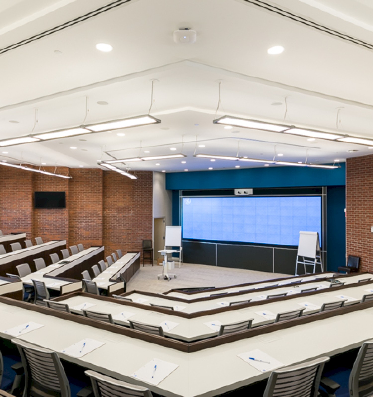 A modern lecture hall with tiered seating, large screen, whiteboards, and well-lit environment, likely for educational purposes.