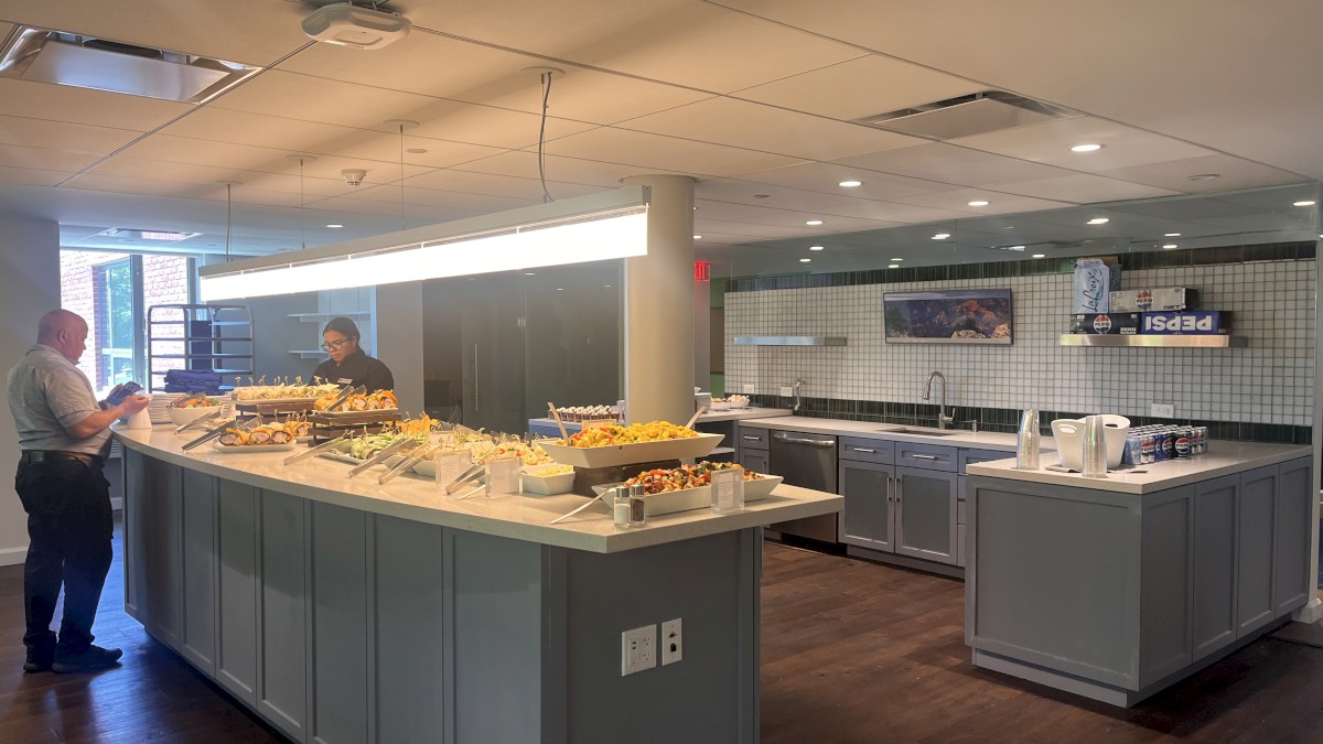 A man looks at his phone while a woman prepares food at a buffet in a modern, well-lit kitchen area.